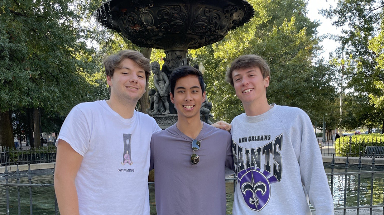 Three young men stand in front of a fountain.