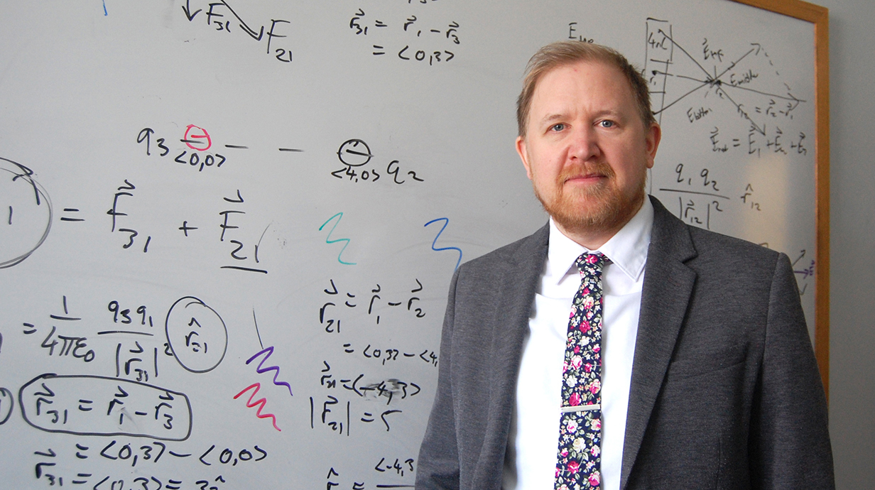 A man wearing a suit stands in front of a whiteboard covered in complex equations.
