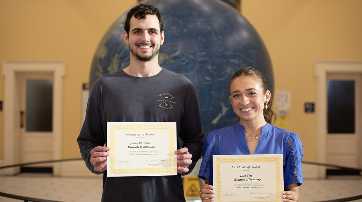 A young man and young woman hold award certificates in front of a giant world globe.