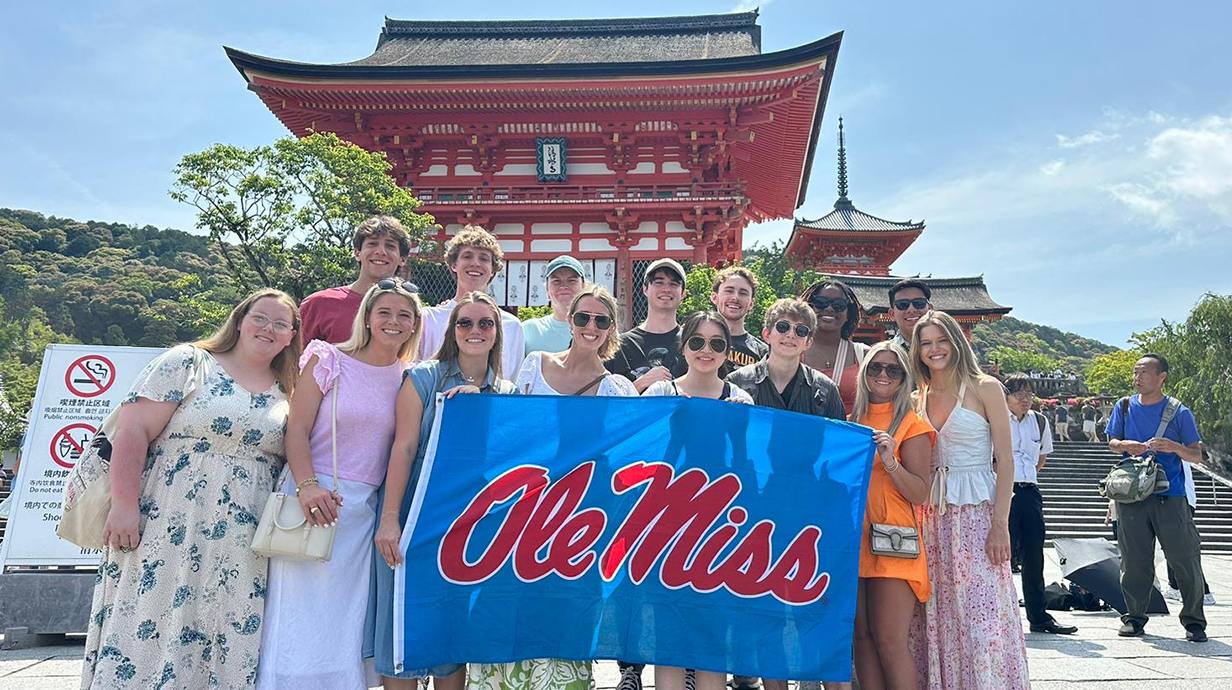 A large group of young people holding a red-and-blue flag that reads 'Ole Miss' stands in front of a Japanese temple.