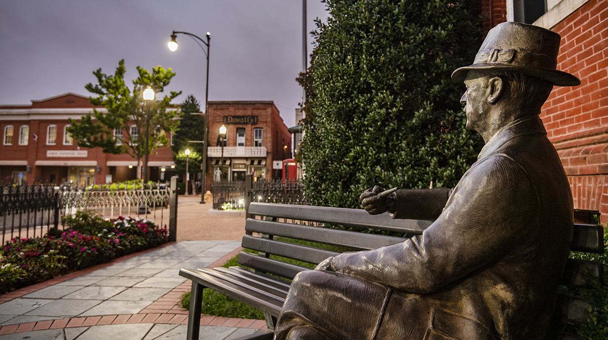 A statue of a man wearing a hat sits on a bench overlooking a town square.