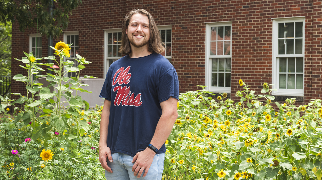 A young man wearing an Ole Miss T-shirt stands in front of a colorful flower garden.