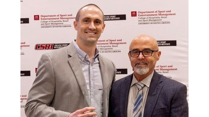 A man presents a crystal award to another man in front of a printed backdrop.