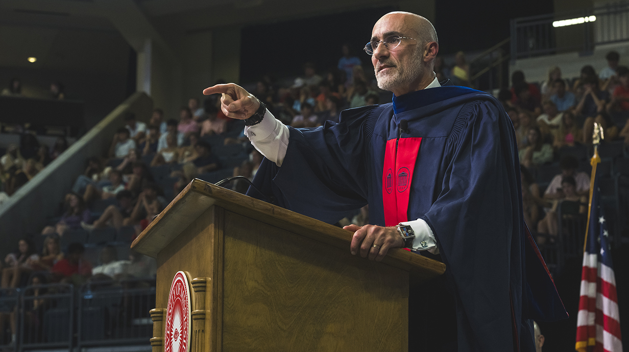 A man wearing a dark blue academic robe speaks to a crowd in an auditorium.