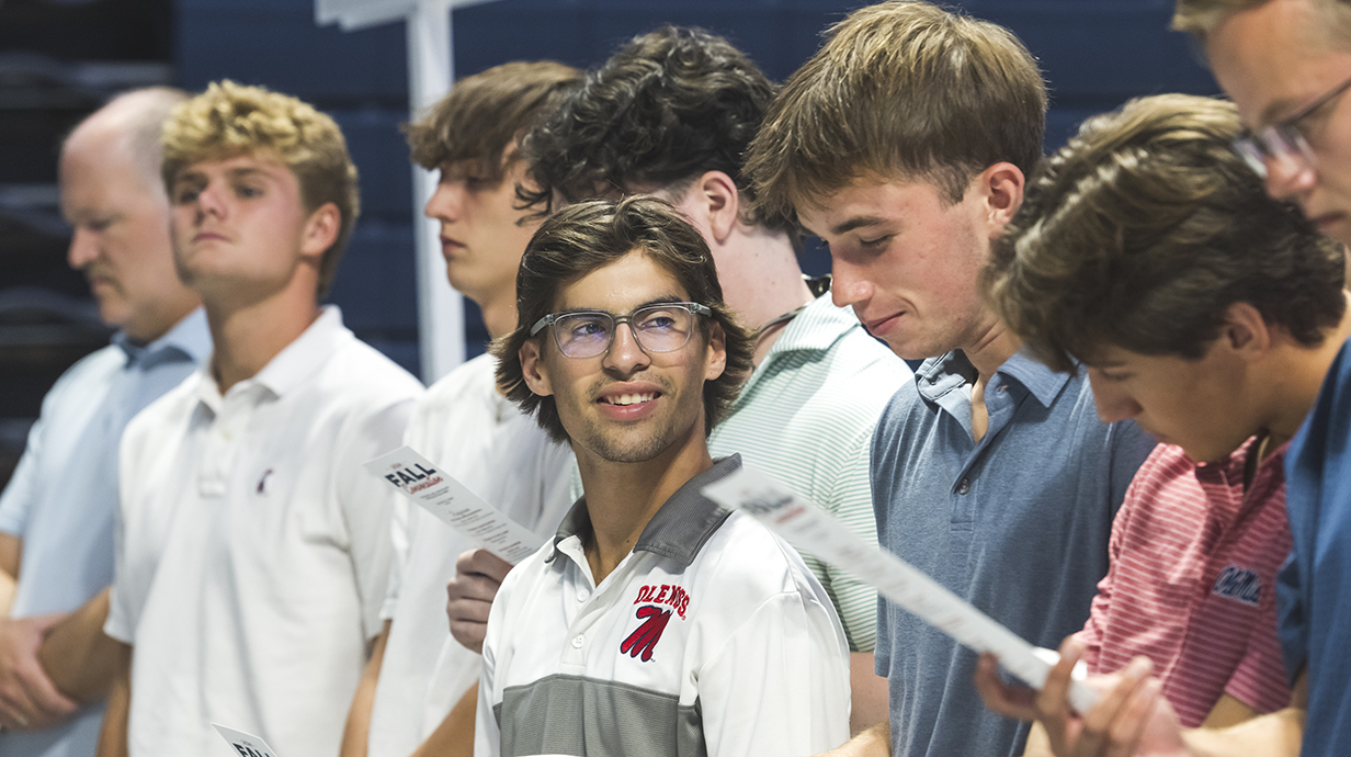 Several young men read from printed programs in a crowded auditorium.