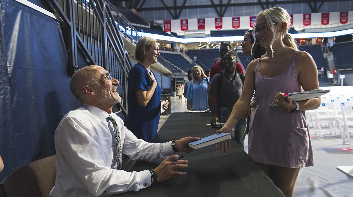 A seated man signs books for a young woman while other people wait in line behind her.