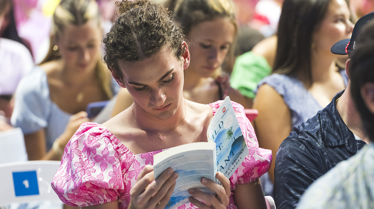 A young man waearing a pink blouse thumbs through a book while seated in a crowded auditorium.