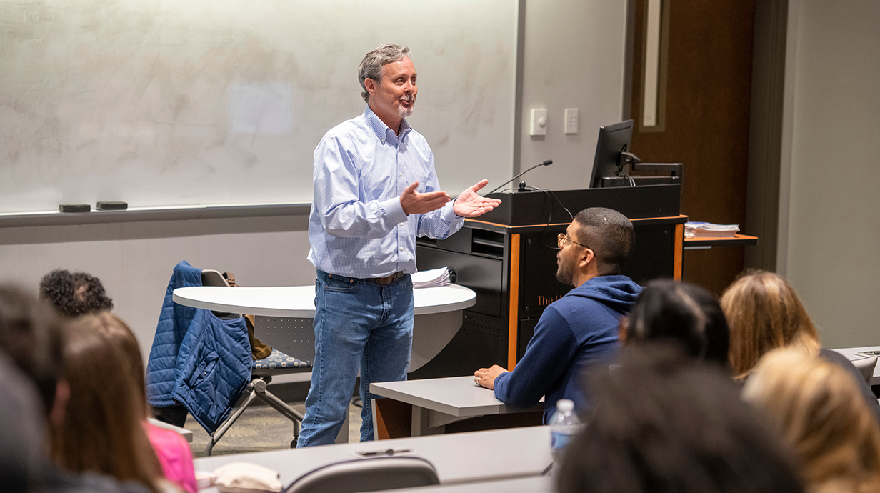 A man standing at a whiteboard lectures to students in a classroom.