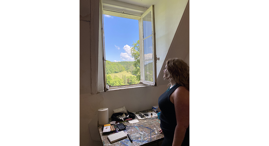 A woman gazes out of a window above an artist's desk.