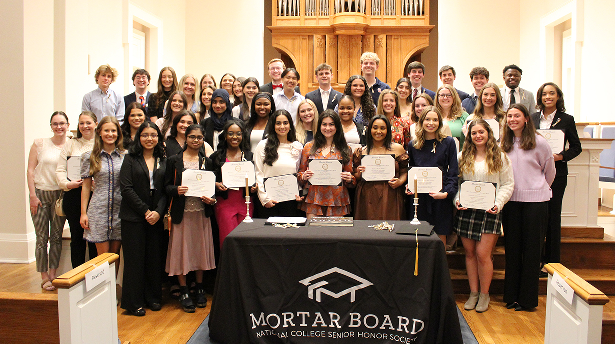 A large group of young people pose for a group photo at the ftont of a chapel behind a Mortar Board honorary banner.