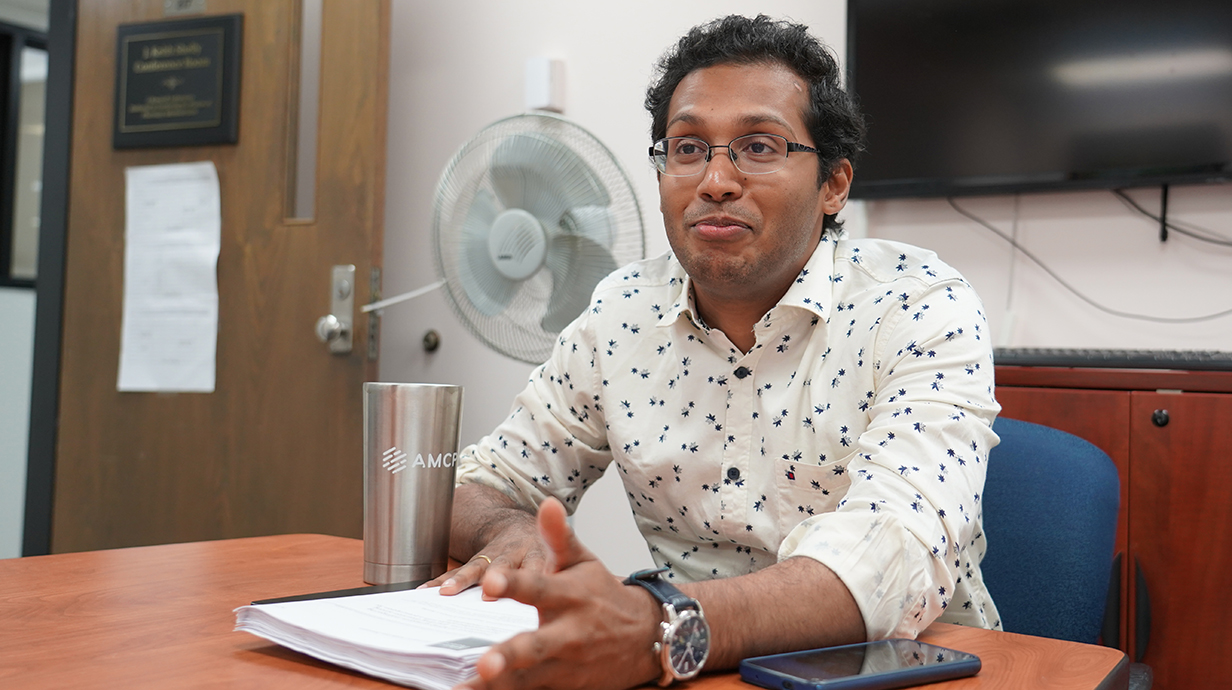 A man sitting at a table in front of a wall-mounted TV gestures to people off-camera.