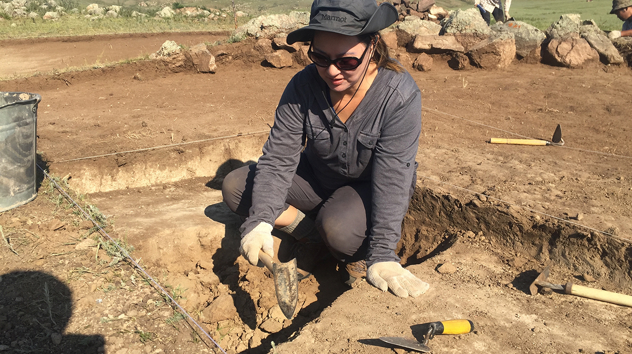 A woman wearing sunglasses and a floppy gray hat uses a small spade to dig at an archaeological site.