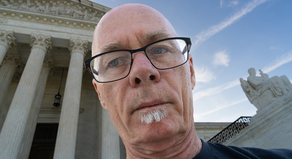 A man takes a close-up selfie in front of a white-columned building.