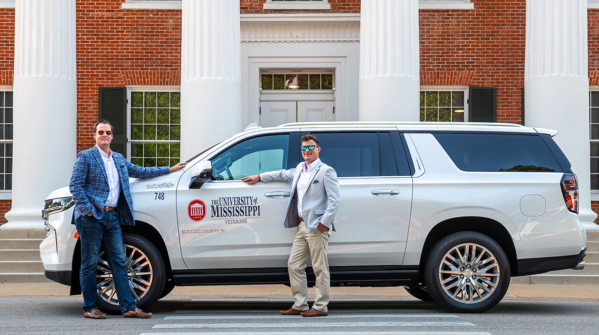 Two men lean against a white SUV in front of a red brick building with large white columns.