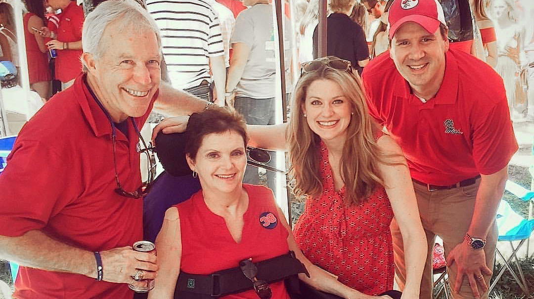 Two men and a woman gather around a woman in a wheelchair, all of them wearing Ole Miss gear, at an outdoor tailgate party.