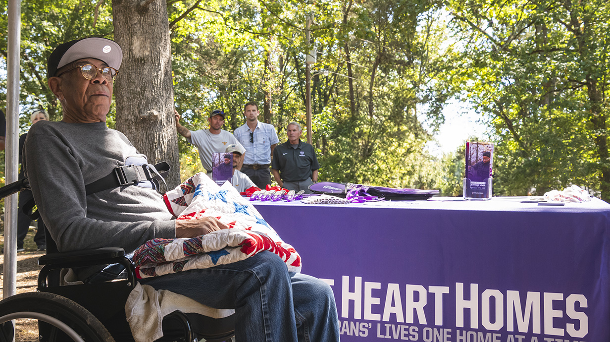 A man sits outdoors in a wheelchair in front of a table with a purple drape reading 'Purple Heart Homes.'