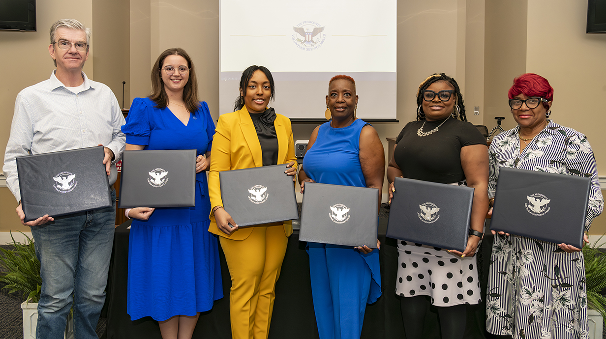 Seven people holding award folders stand in front of a projection screen.