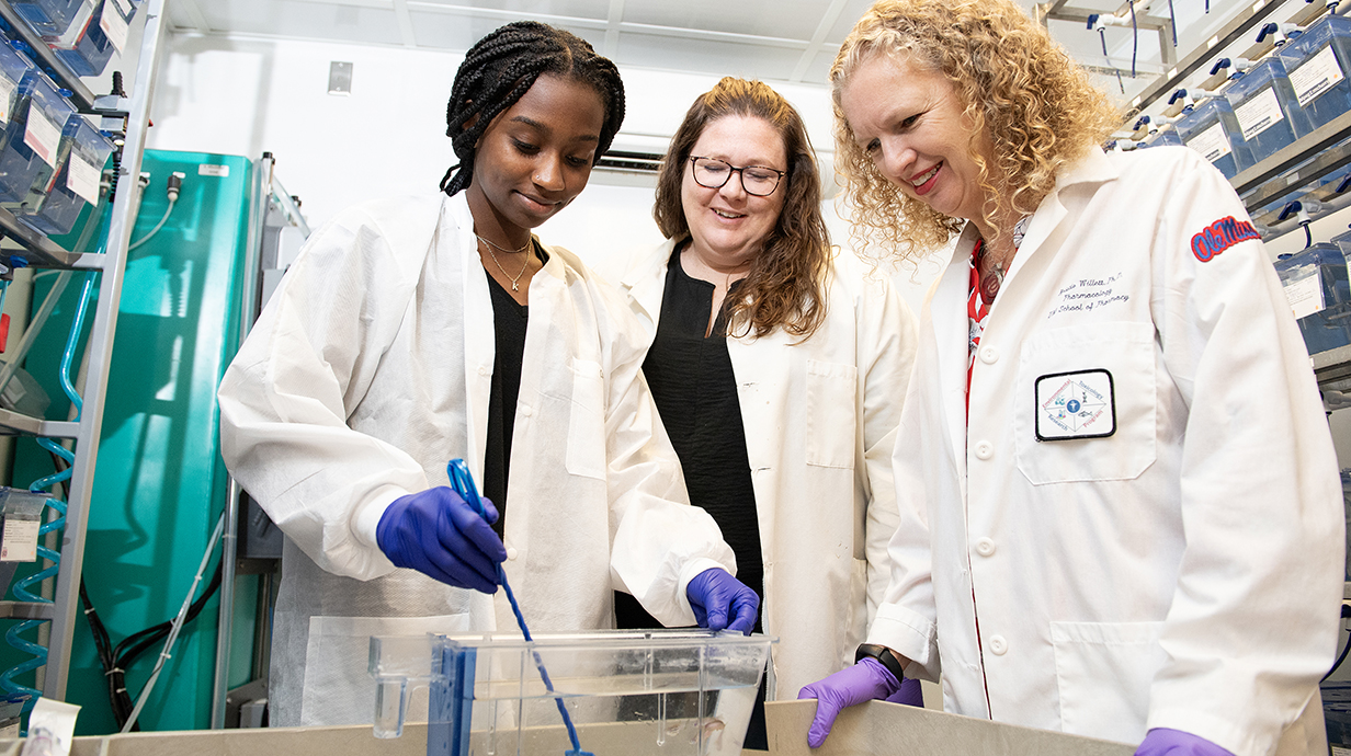 Two women wearing white lab coats watch as a young woman uses a small net to retrieve a minnow from a fishtank in a laboratory.