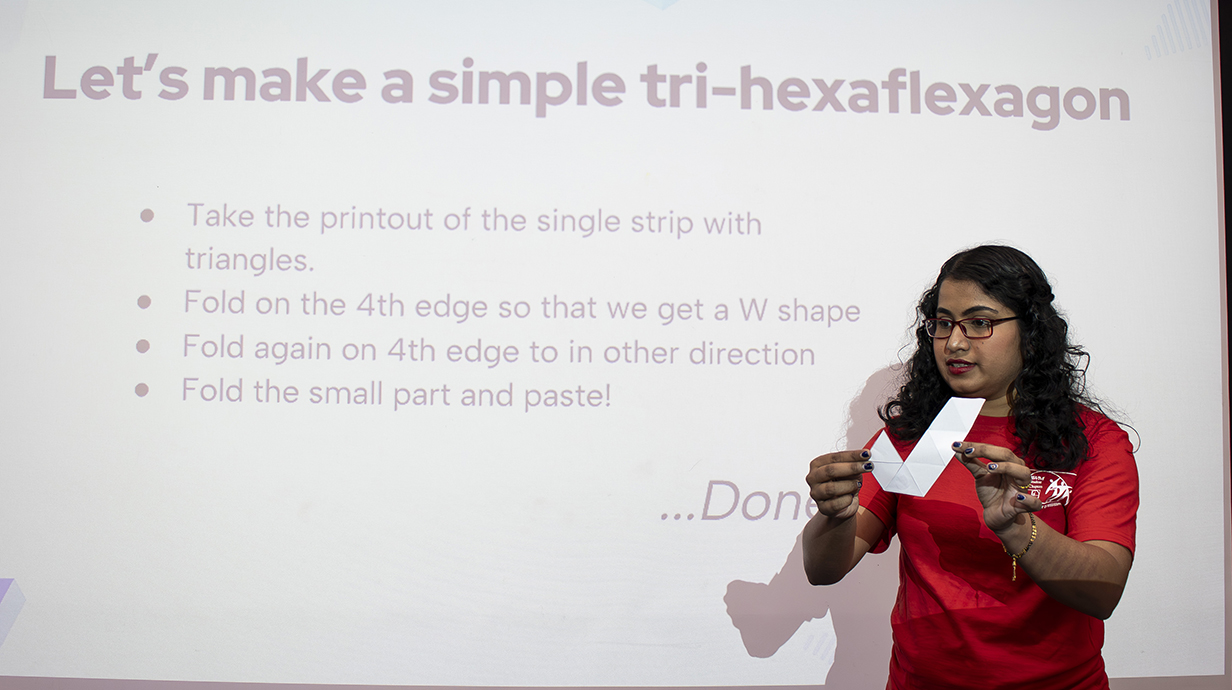 A young woman standing in front of a projection screen uses a strip of paper to demonstrate a mathematical concept.