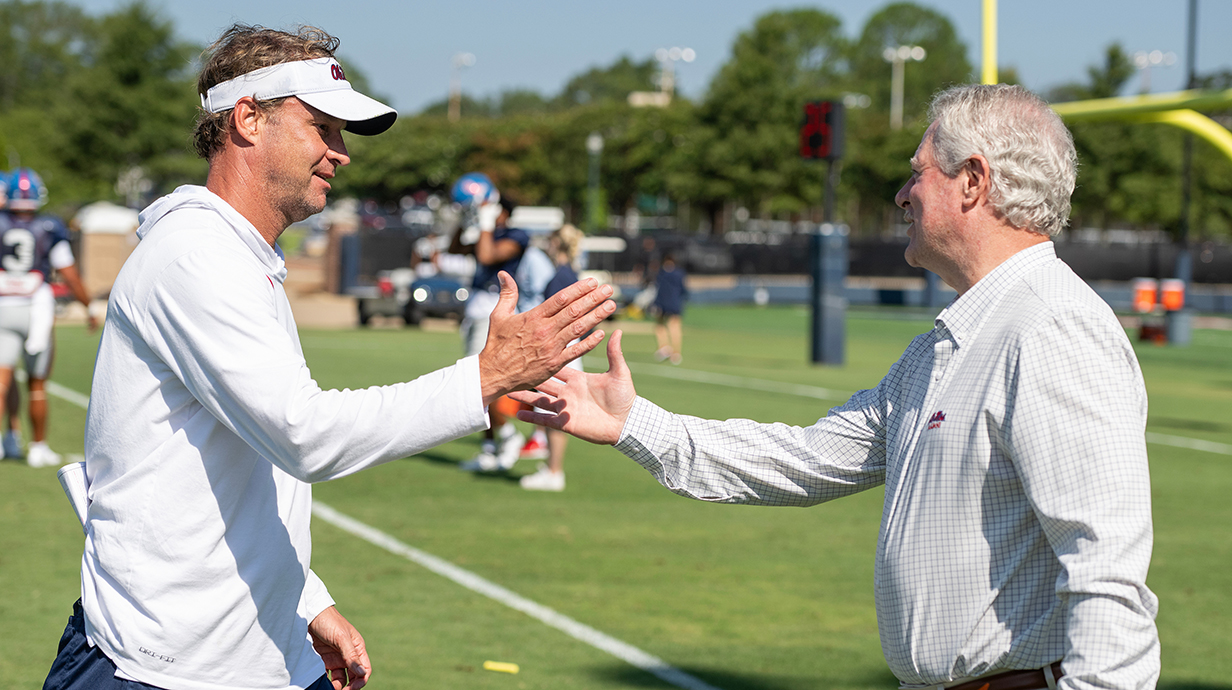 Two men shake hands standing near a football field.