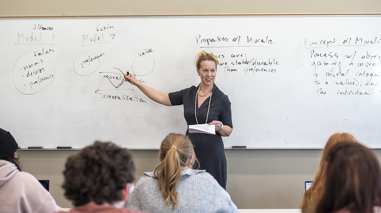 A woman points to writing on a whiteboard in front of a classroom full of students.