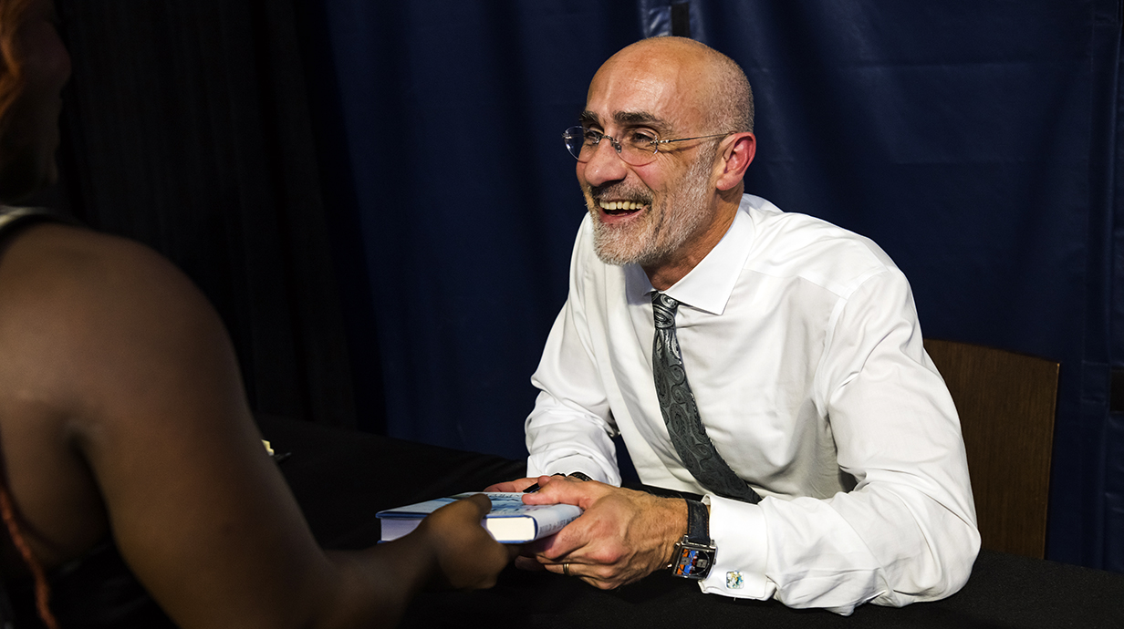 A seated man wearing a tie and holding a book smiles at a person off-camera.