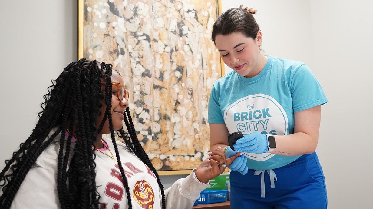 A young woman helps another woman check her blood sugar using a fingertip device at a pharmacy.