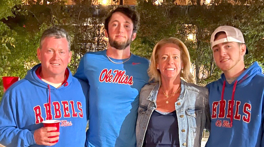 A family of four, three of them wearing Ole Miss athletics gear, stand on an outside porch.