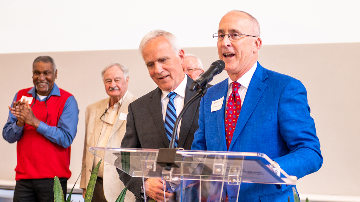 A man in a suit speaks at a microphone and a man beside him bows his head as people around them applaud.