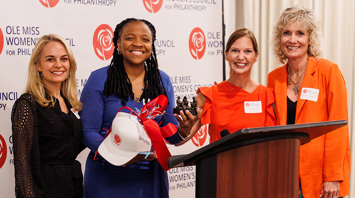 Four women stand smiling in front of a backdrop reading 'Ole Miss Women's Council for Philanthropy.' One holds a hat, red-and-blue ribbons and a small statue.