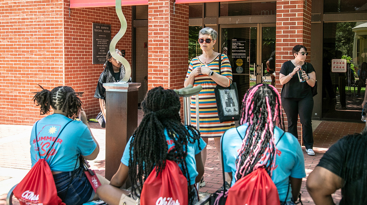 A woman standing beside a piece of sculpture talks to a group of young people outside a red brick building.