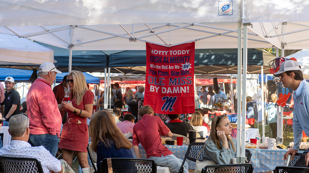 Football fans chat at a pregame tailgate underneath a pop-up tent.