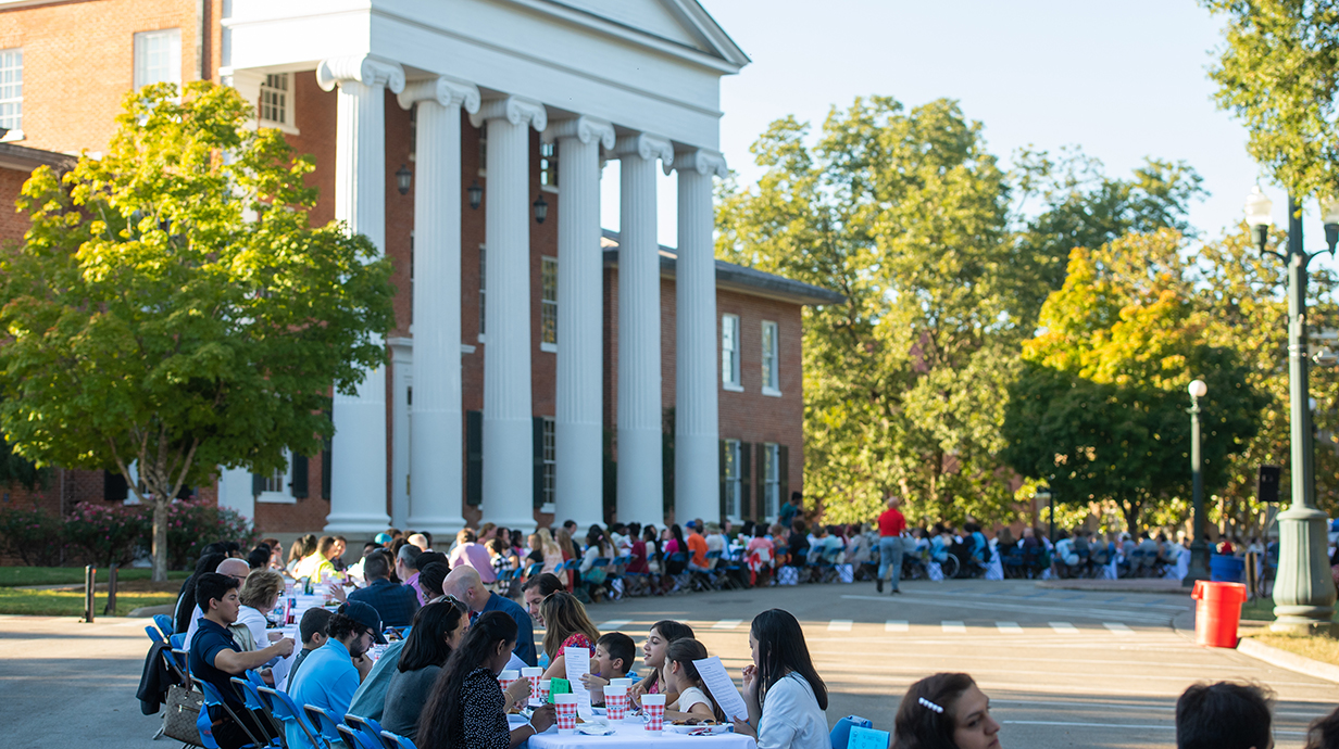 A large group of people sit at a long line of tables set up on a street outside a white-columned building on a sunny day.