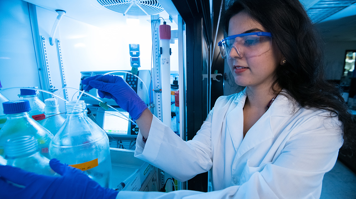 A woman wearing a white lab coat and safety glasses works in a laboratory.