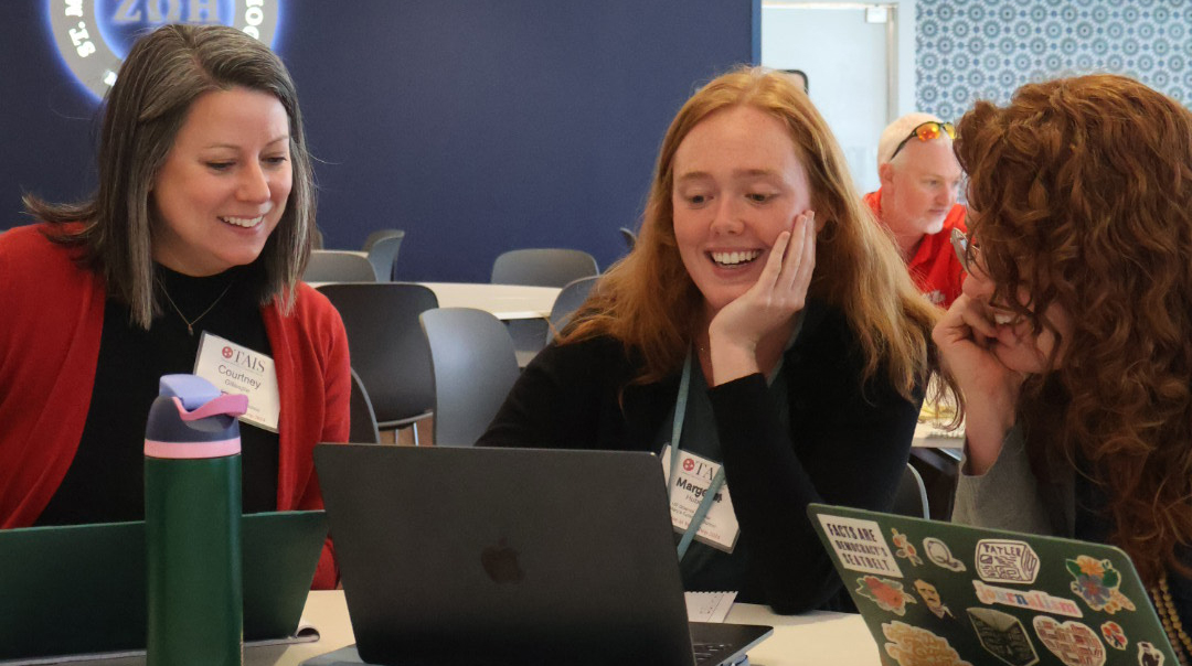 Three women sitting around a table in a conference hall smile as they read off a laptop screen in front of the middle woman.