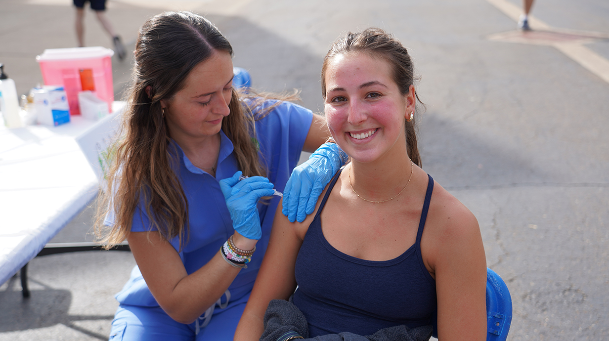 A young woman wearing medical scrubs gives another young woman a glu shot at an outdoor immunization drive.