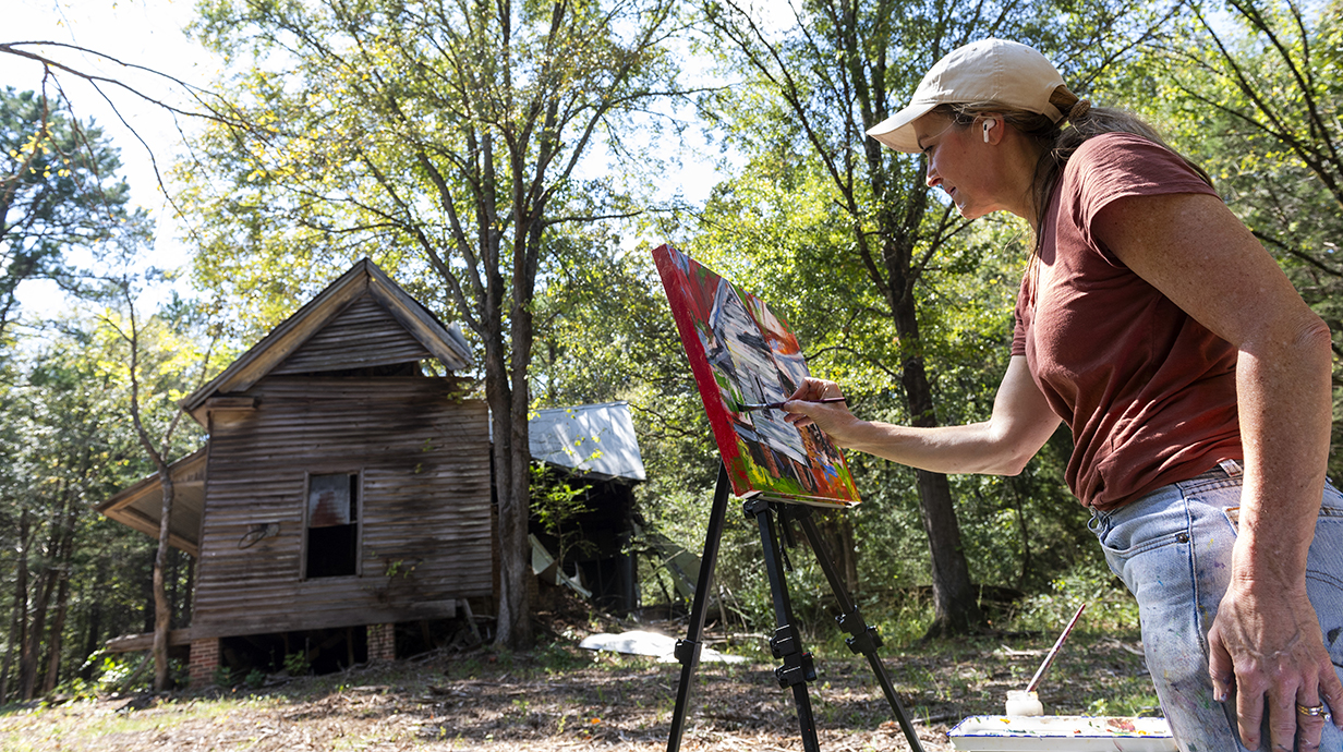 A woman works on a painting outdoors, near a dilapidated old building in a wooded area.