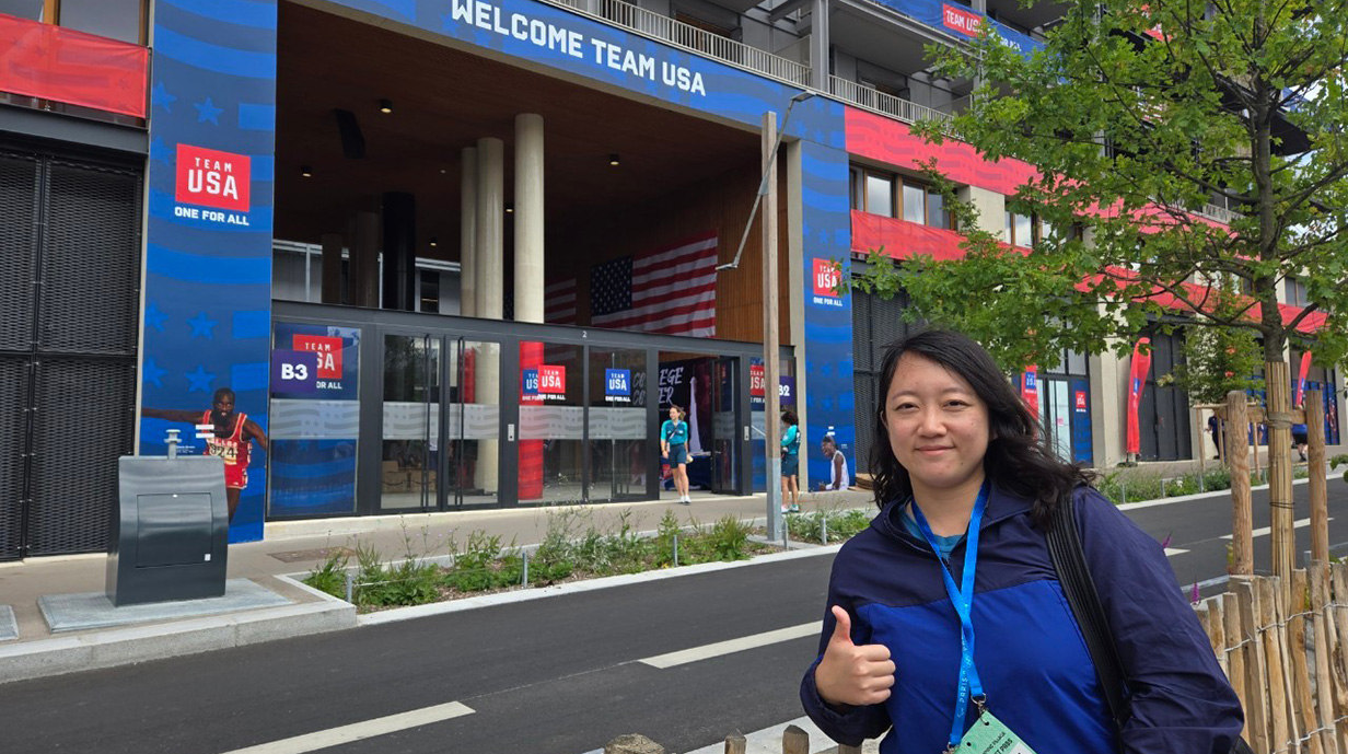 A woman stands across a street from a building with a huge blue banner reading 'Welcome Team USA'