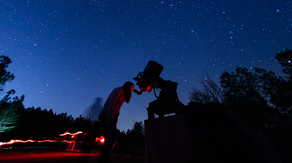 A young woman looks through a telescope against a darkened sky.