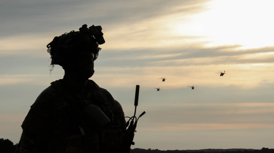 A silhouetted soldier watches as four helicoptors fly against a hazy sky.