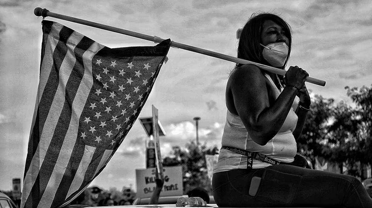 A black-and-white photograph of a woman sitting with an American flag draped behind her.