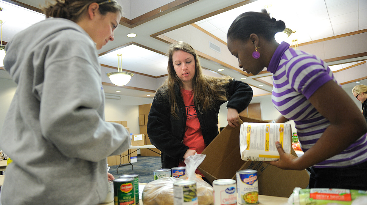 Three young women sort grocery items into a cardboard box.