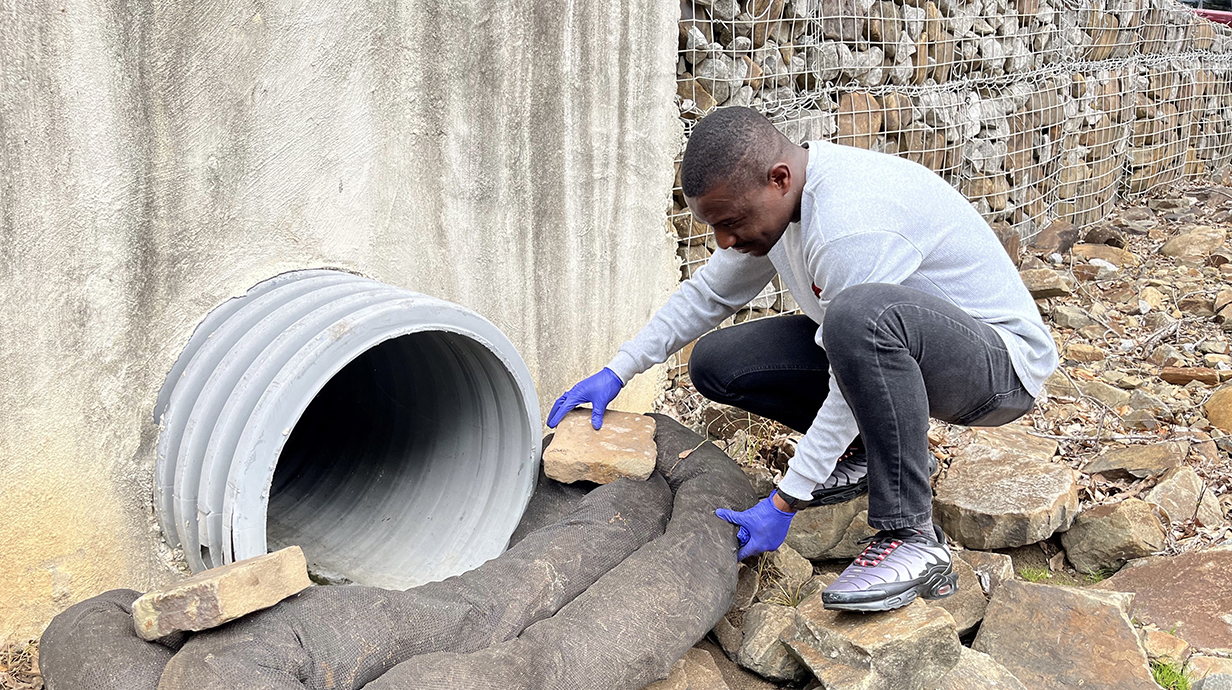 A young man places sandbags on a rock bed beneath a drainage pipe.