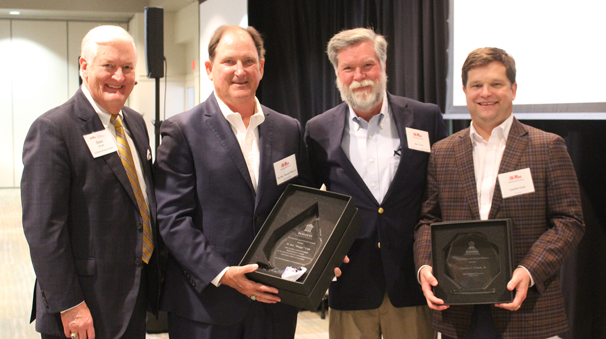 Four men -- two of them holding award presentations -- stand in front of a projection screen in a banquet hall.