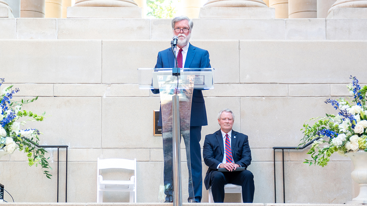 A man wearing a suit speaks at a lectern while another man sits behind him.