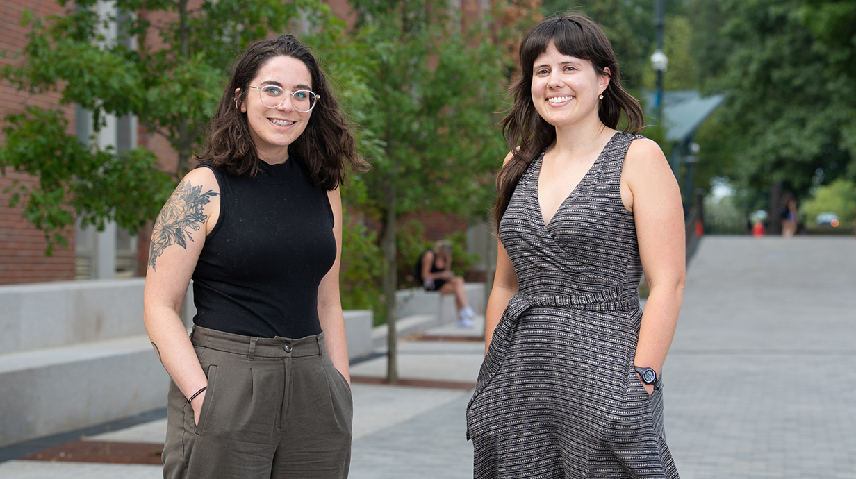 Two women stand on a tree-lined plaza outside a red brick building.