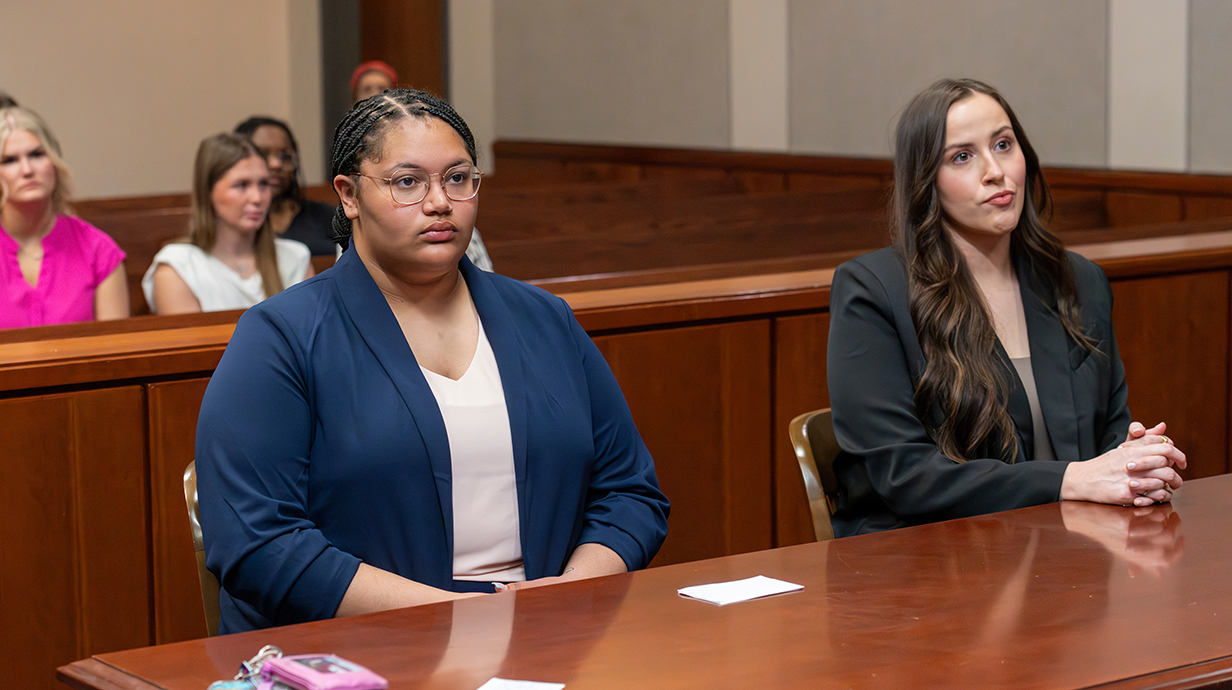 Two young women sit at a table at the front of a courtroom.