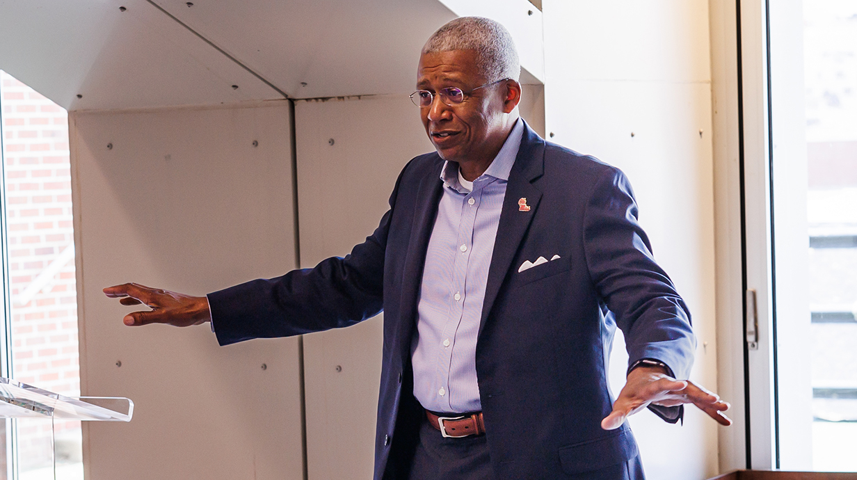 A man in a blue suit gestures while talking to a group of people.