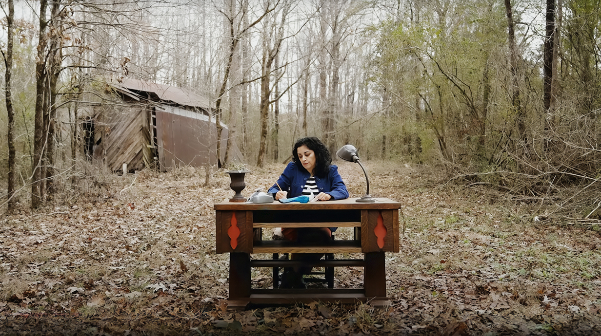 A woman sits at a desk in a clearing in front of trees and a dilapidated building.