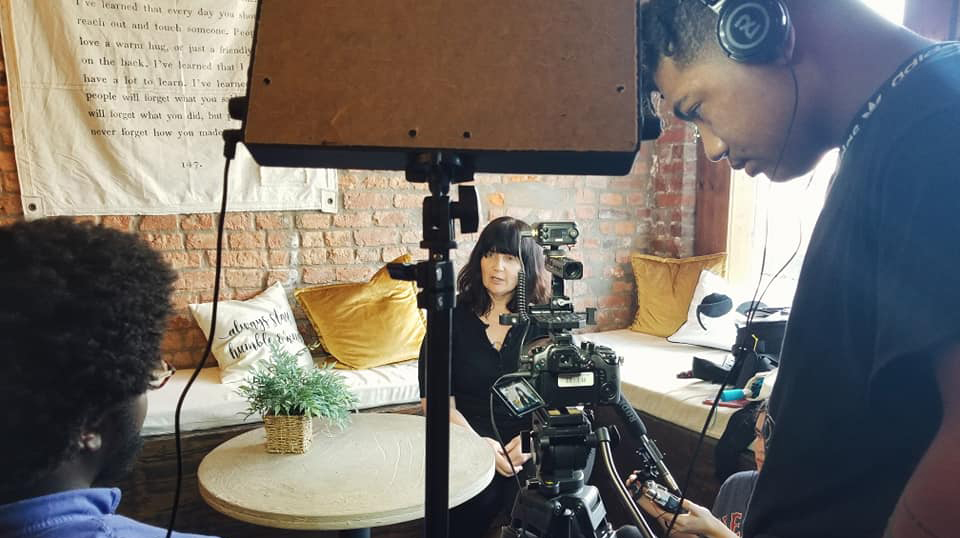 A young man and young woman use a video camera to interview a woman seated on a sofa.